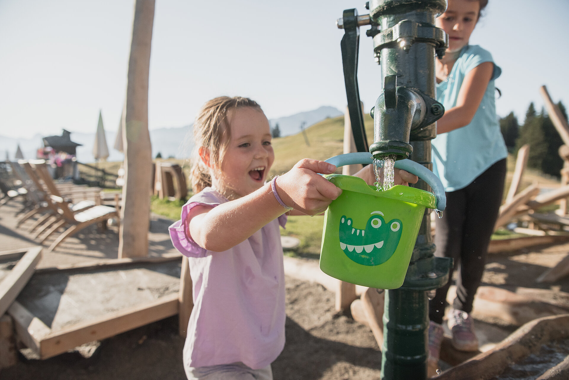 <p>Ein Mädchen im rosa Shirt lässt sich von einer Spielkameradin ihren grünen Kessel mit Wasser am Pumpbrunnen füllen.</p>