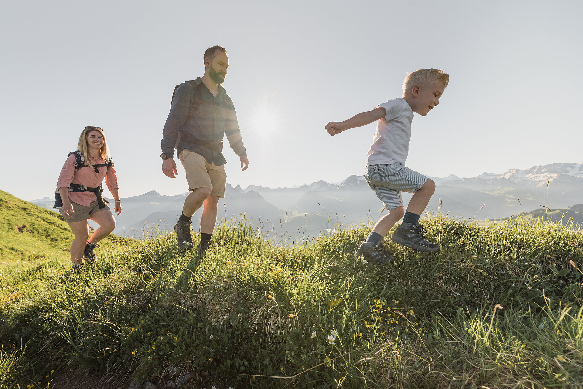 <p>Familie bestehend aus drei Personen beim Wandern über grüne Wiesen bei sommerlichem Wetter.</p>